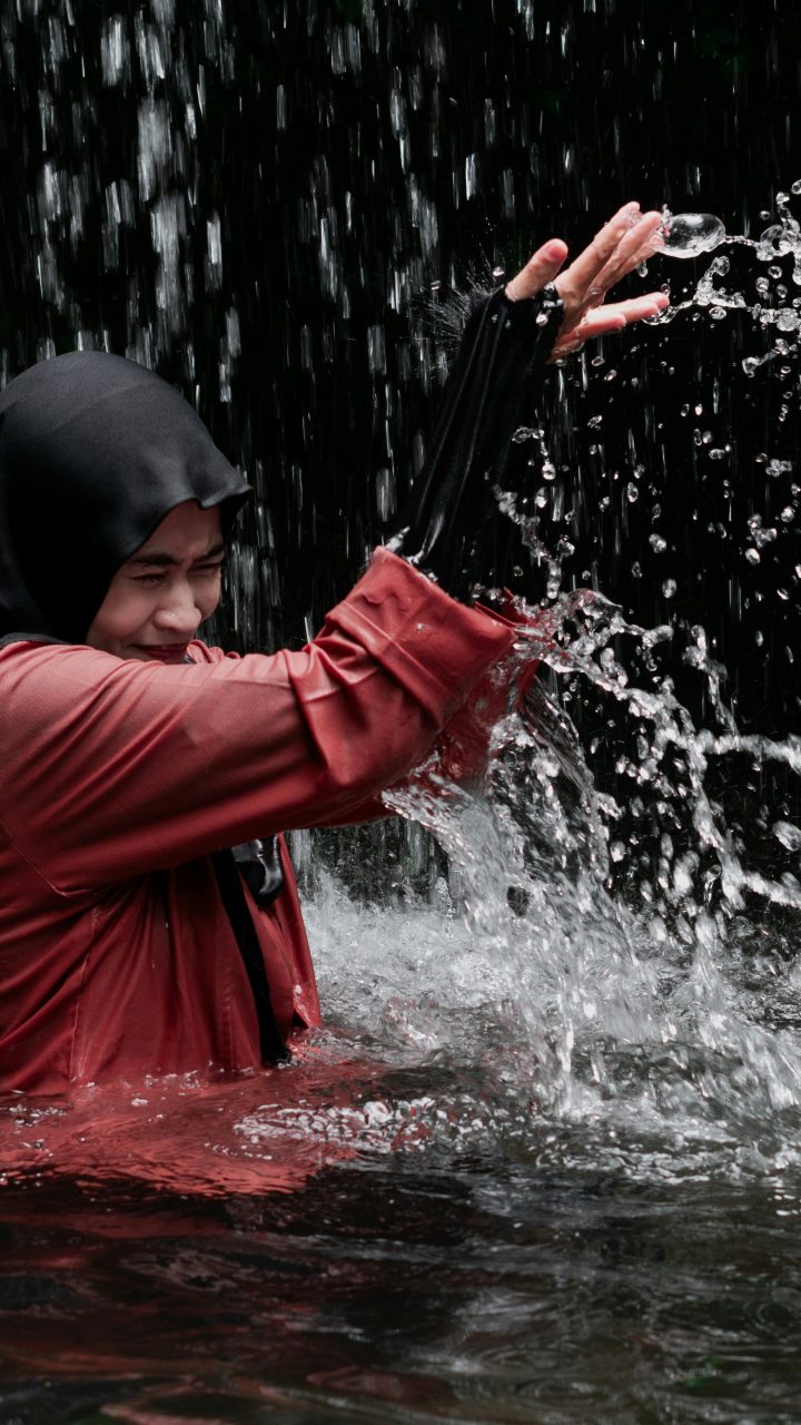 A person in a hijab and red shirt splashing water in a waterfall