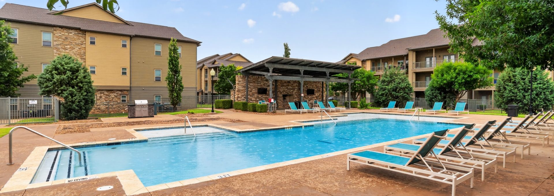 Modern apartment complex pool. The large blue pool, many lounge chairs, outdoor kitchen, & shaded seating. Tan buildings are surrounded by landscaping.