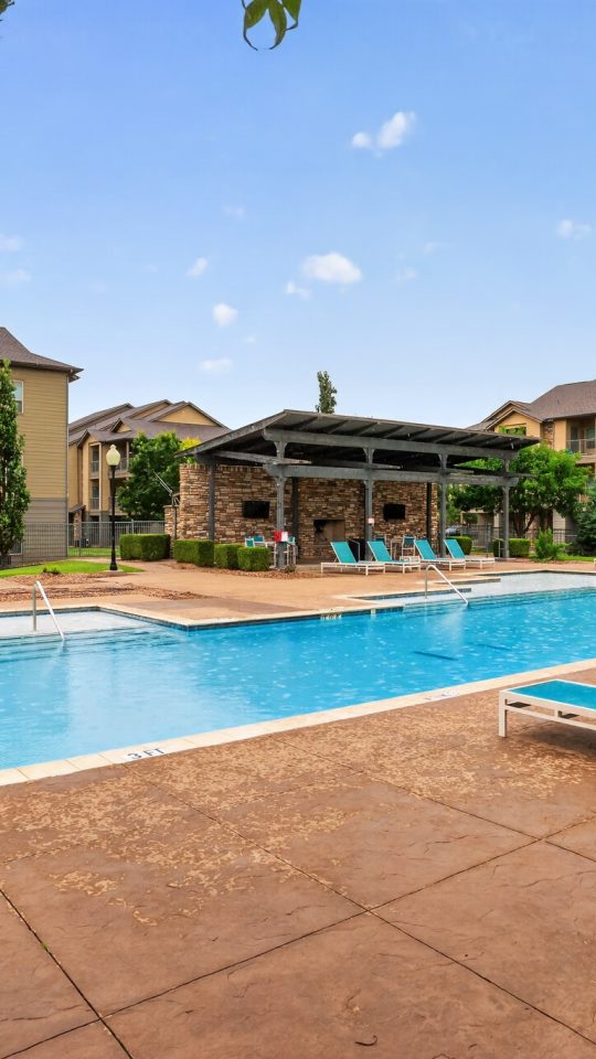Modern apartment complex pool. The large blue pool, many lounge chairs, outdoor kitchen, & shaded seating. Tan buildings are surrounded by landscaping.