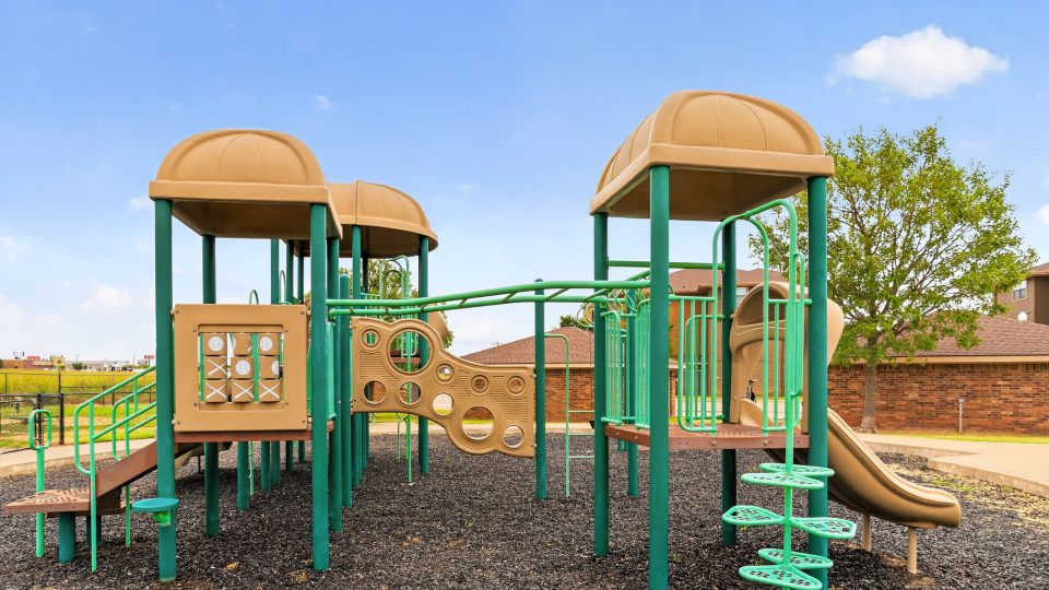 A large community playground has green and tan equipment: slides, monkey bars, and steps on black rubber mulch. Nearby apartment buildings are visible.