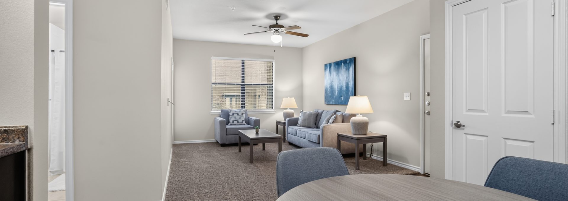 Spacious open-concept apartment dining room flows into a carpeted living area with a blue sofa, armchairs, and a window. Light walls & modern, clean decor.
