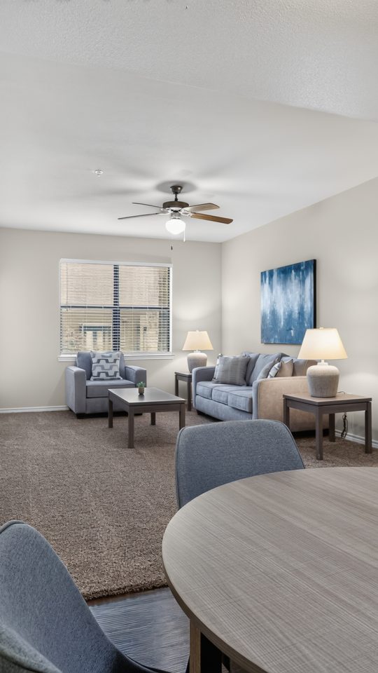 Spacious open-concept apartment dining room flows into a carpeted living area with a blue sofa, armchairs, and a window. Light walls & modern, clean decor.