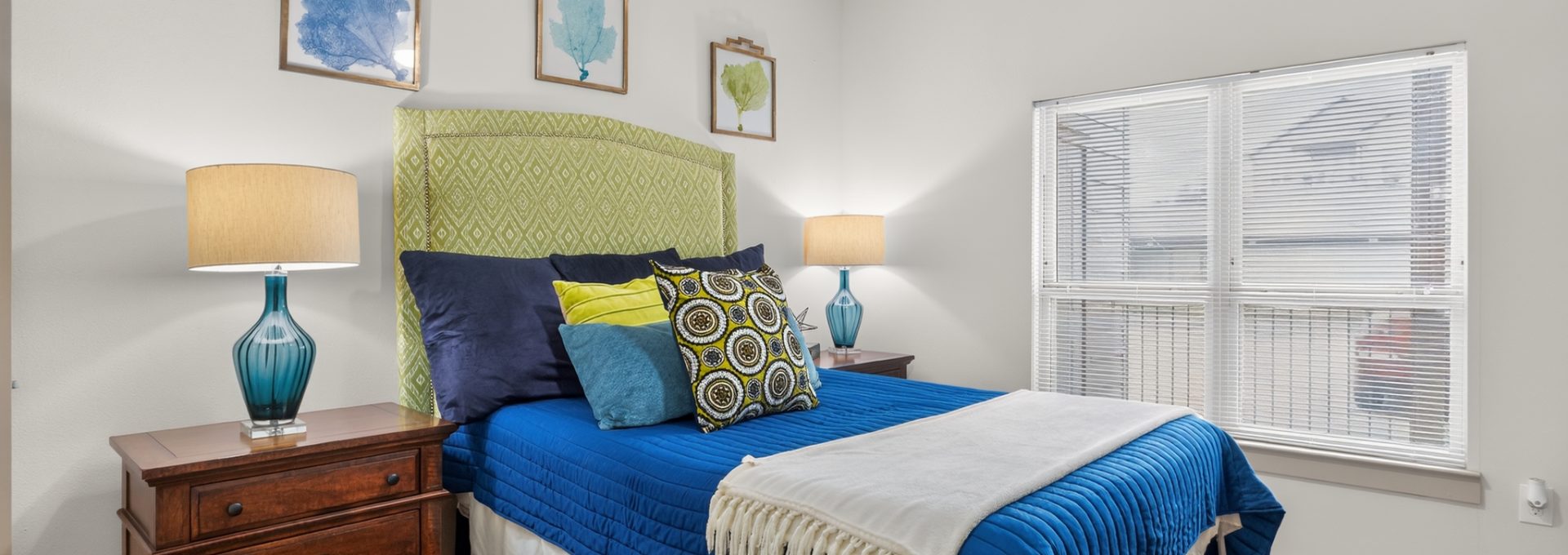 A cozy bedroom with a bed, green headboard, blue comforter, and pillows. Nightstands with lamps, a window with blinds, brown carpet, and a ceiling fan.