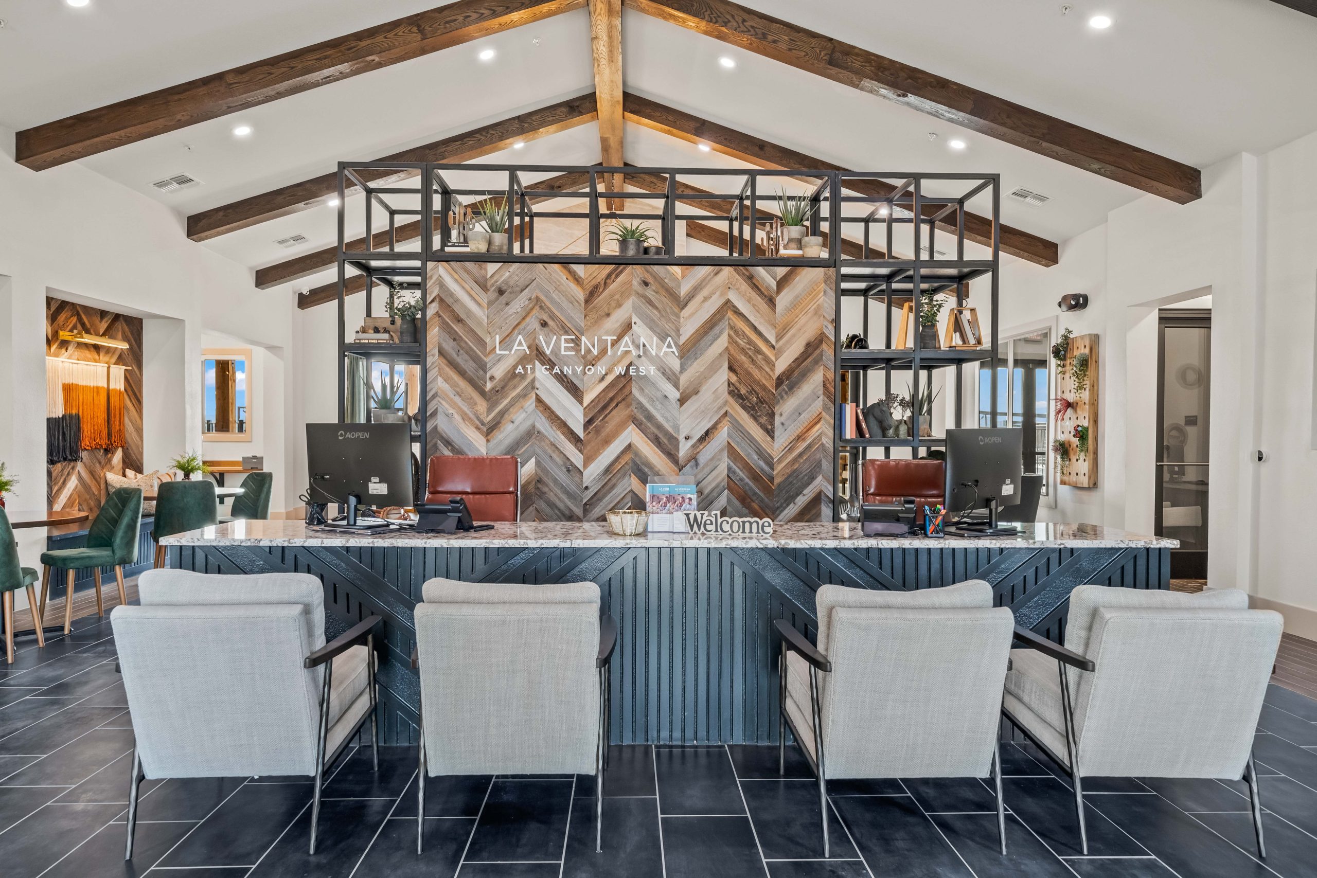 A bright, modern leasing office at La Ventana at Canyon West featuring vaulted ceilings with exposed wooden beams, a large reception desk, contemporary seating, and a wood-paneled accent wall displaying the La Ventana logo