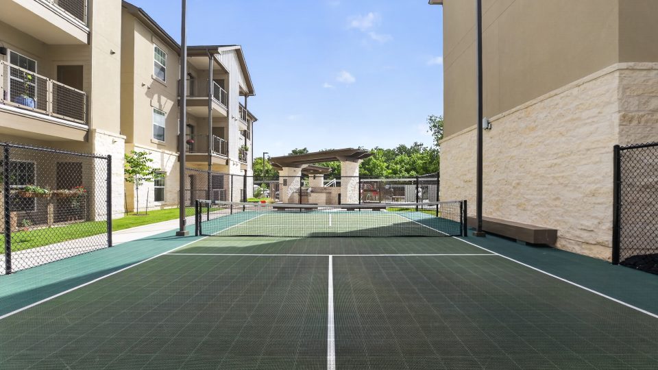 Apartment complex features a green tennis/pickleball court. Tan buildings with balconies flank the court, with a covered communal grilling pavilion behind.