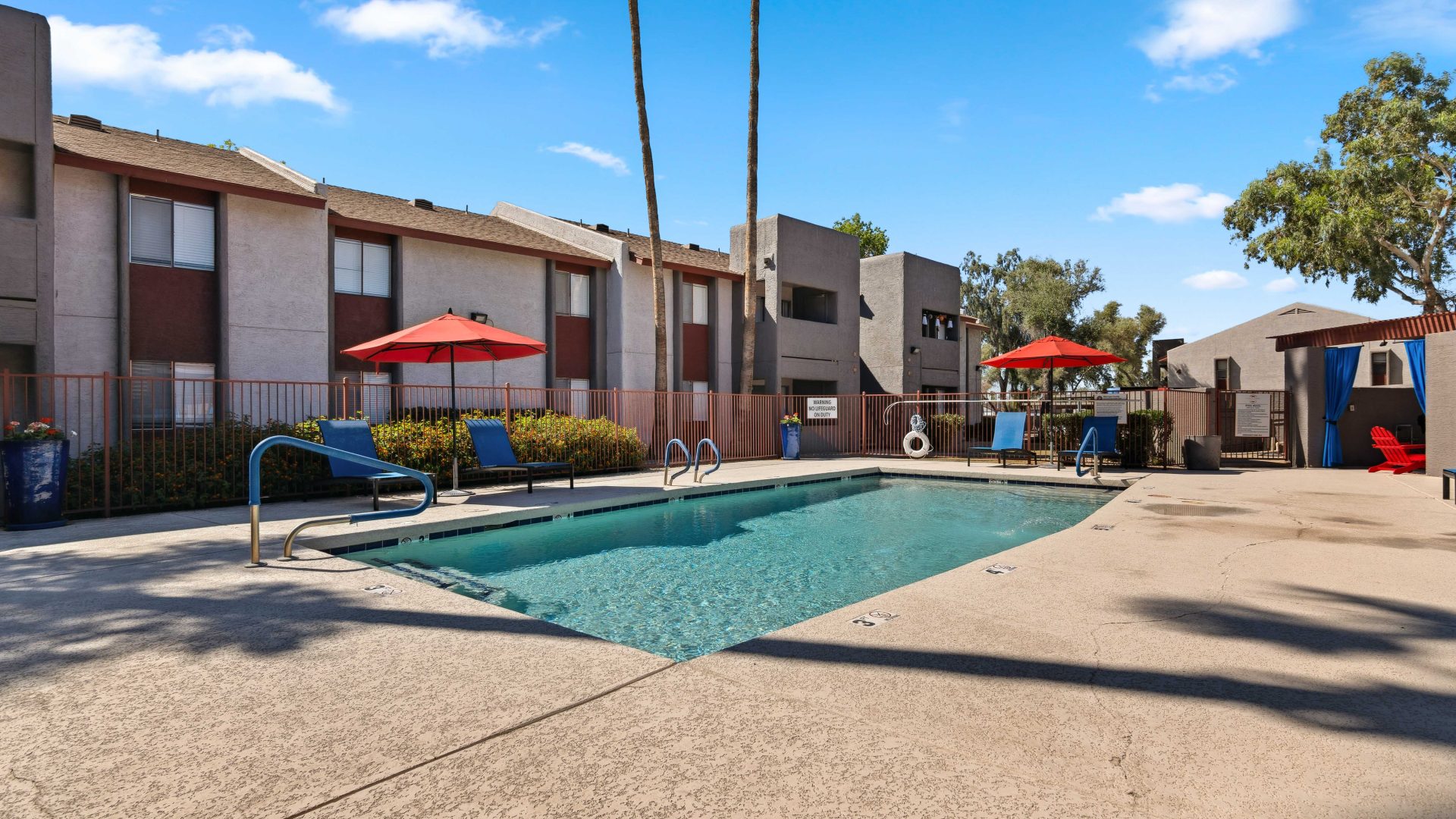 Resort-style swimming pool with red umbrellas, lounge seating, and palm trees at Country Gables in Glendale, AZ