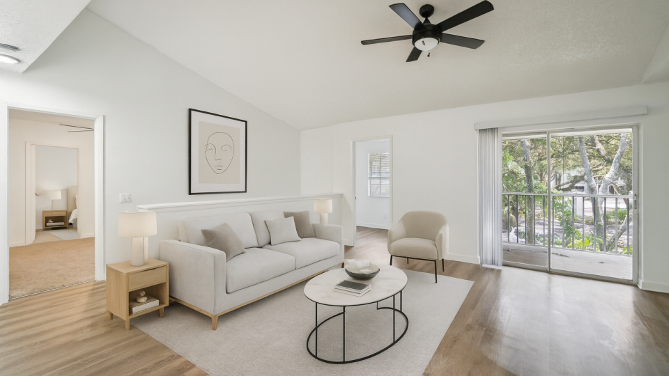 A serene and modern living room features a plush, light-colored sofa adorned with throw pillows, positioned centrally on a large area rug. The room is accented with a contemporary coffee table holding a few decorative items, and a stylish side table with a lamp. A comfortable armchair sits nearby, and a large framed artwork hangs above the sofa. The space is brightened by natural light streaming through a sliding glass door leading to a balcony, creating a tranquil atmosphere perfect for relaxation. The room's minimalist design and neutral color palette enhance its inviting and cozy ambiance.