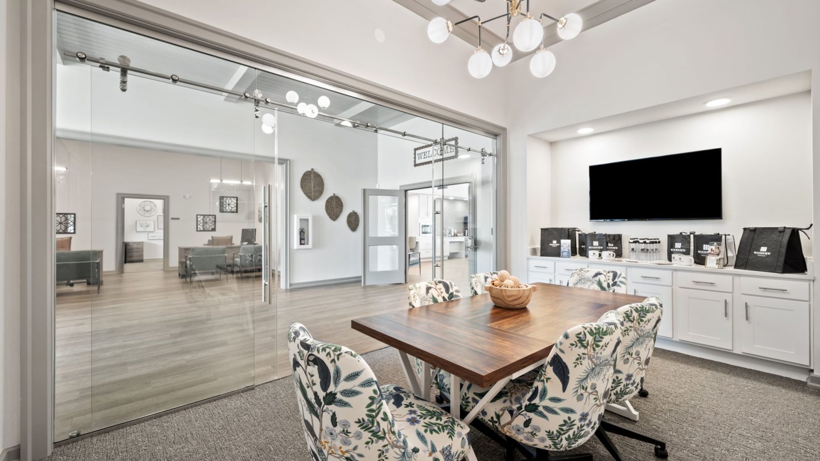 Modern leasing office with a large wooden meeting table, floral chairs, TV, and cabinets. Glass doors open to a lobby.