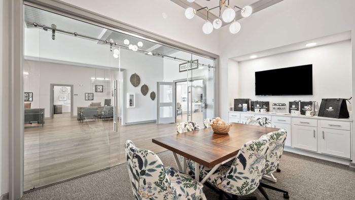 Modern leasing office with a large wooden meeting table, floral chairs, TV, and cabinets. Glass doors open to a lobby.