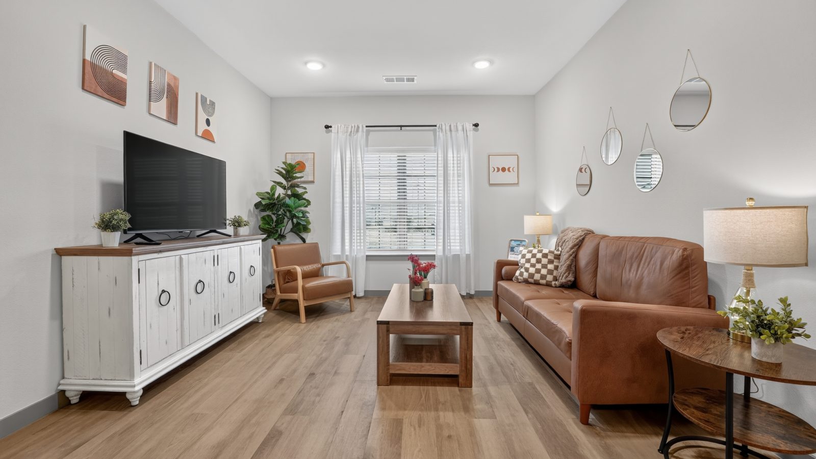 Bright, furnished living room with light wood floors. Features a brown leather sofa, armchair, coffee table, and TV.