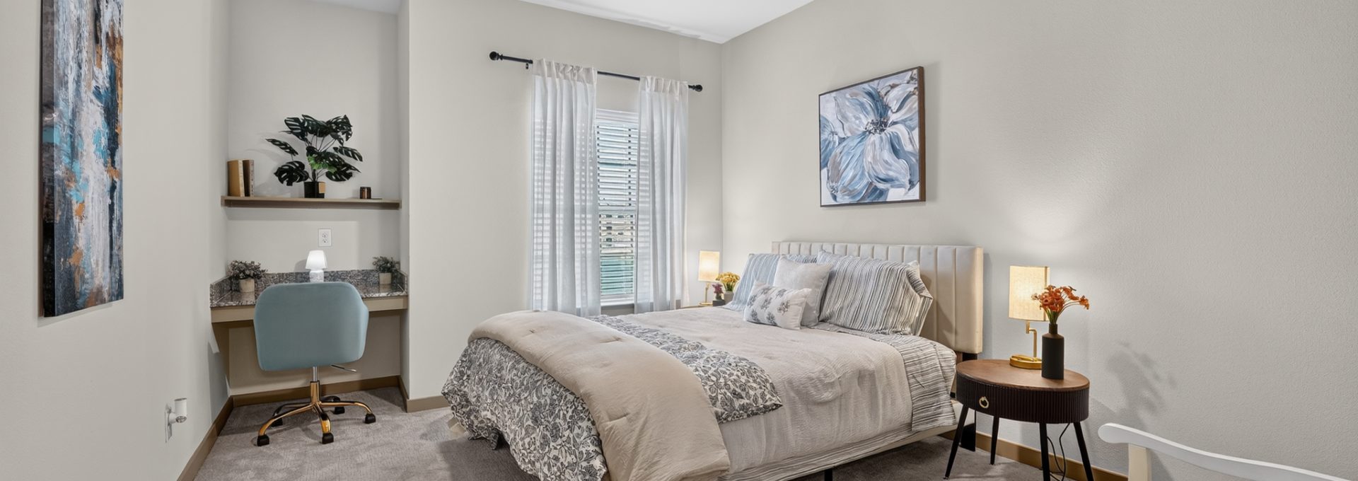 Bright, modern apartment bedroom with light walls, soft grey carpet, a comfy bed, built-in desk, and ceiling fan.
