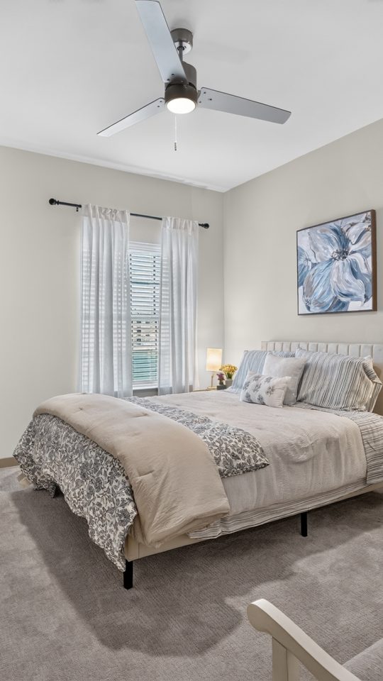 Bright, modern apartment bedroom with light walls, soft grey carpet, a comfy bed, built-in desk, and ceiling fan.