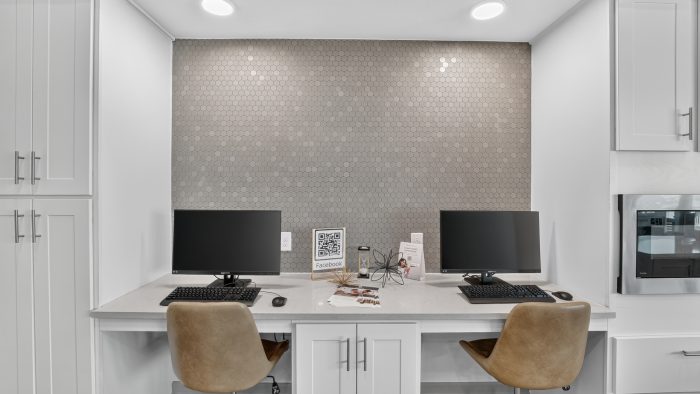 Community business center with two computer stations. White cabinets, light gray hexagonal tile wall, and comfy brown chairs.