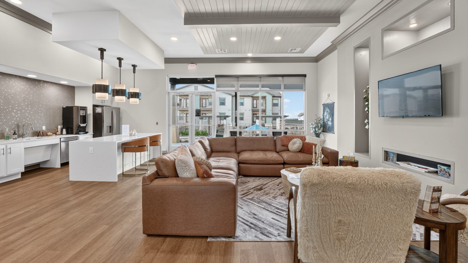 Modern, open common area with kitchen, island seating, brown sectional sofa, TV, and large windows to other units.