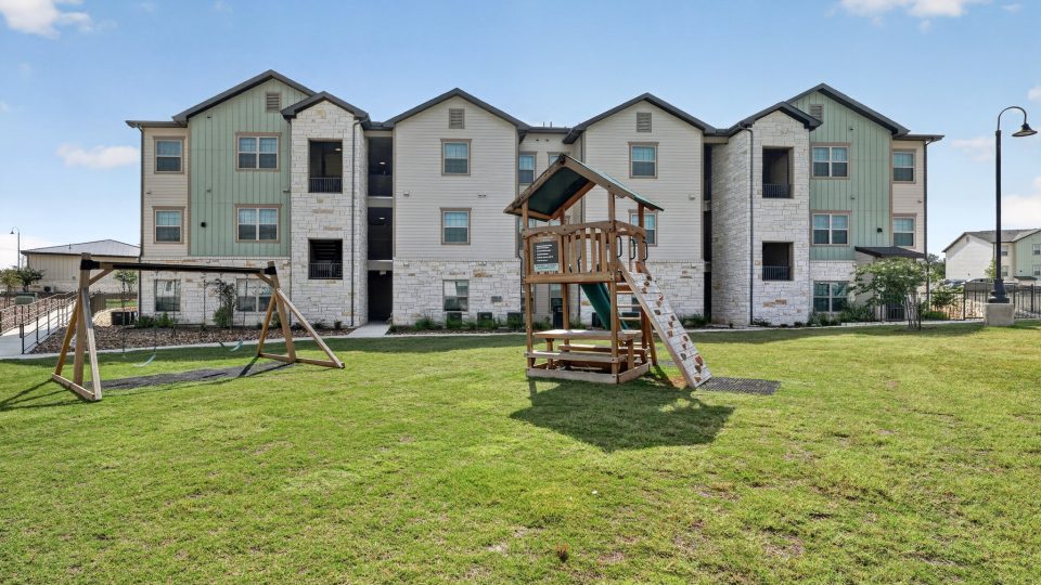 Modern three-story apartment buildings with stone & green siding. A large grassy common area has a swing set & playset.