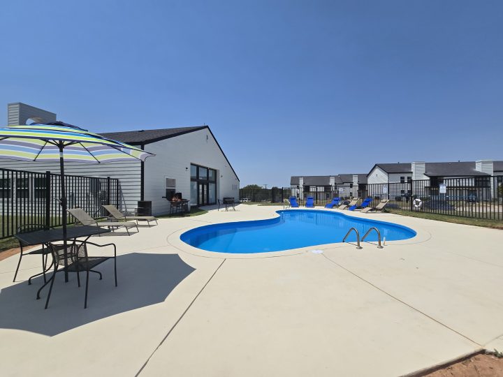 A serene outdoor scene featuring a sparkling blue swimming pool as the centerpiece, surrounded by a spacious patio with comfortable lounge chairs and a colorful umbrella. The area is enclosed by a black metal fence, providing a sense of privacy and security. The setting is enhanced by a modern white building with large windows, contributing to a tranquil and inviting atmosphere perfect for relaxation and leisure. The overall ambiance suggests a peaceful retreat ideal for enjoying sunny days and unwinding by the poolside.