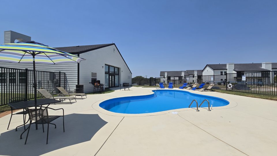A serene outdoor scene featuring a sparkling blue swimming pool as the centerpiece, surrounded by a spacious patio with comfortable lounge chairs and a colorful umbrella. The area is enclosed by a black metal fence, providing a sense of privacy and security. The setting is enhanced by a modern white building with large windows, contributing to a tranquil and inviting atmosphere perfect for relaxation and leisure. The overall ambiance suggests a peaceful retreat ideal for enjoying sunny days and unwinding by the poolside.