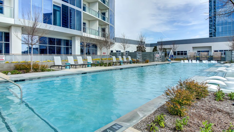 Modern glass high-rise apartment overlooks a bright blue outdoor pool. The pool area features lounge chairs, water fountains, and minimal landscaping.