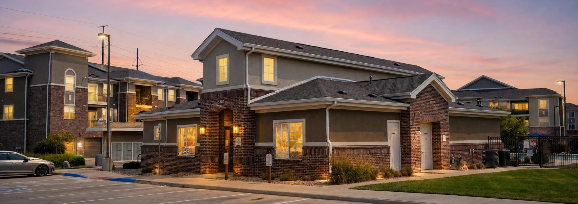 Outlook Blvd apartments. Modern brick clubhouse and buildings at sunset. Lit windows, empty parking lot.