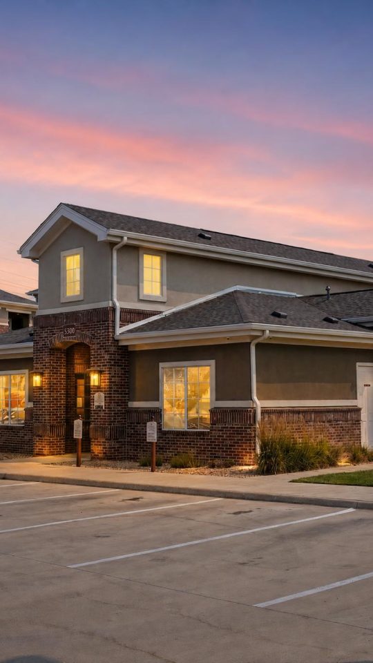 Outlook Blvd apartments. Modern brick clubhouse and buildings at sunset. Lit windows, empty parking lot.