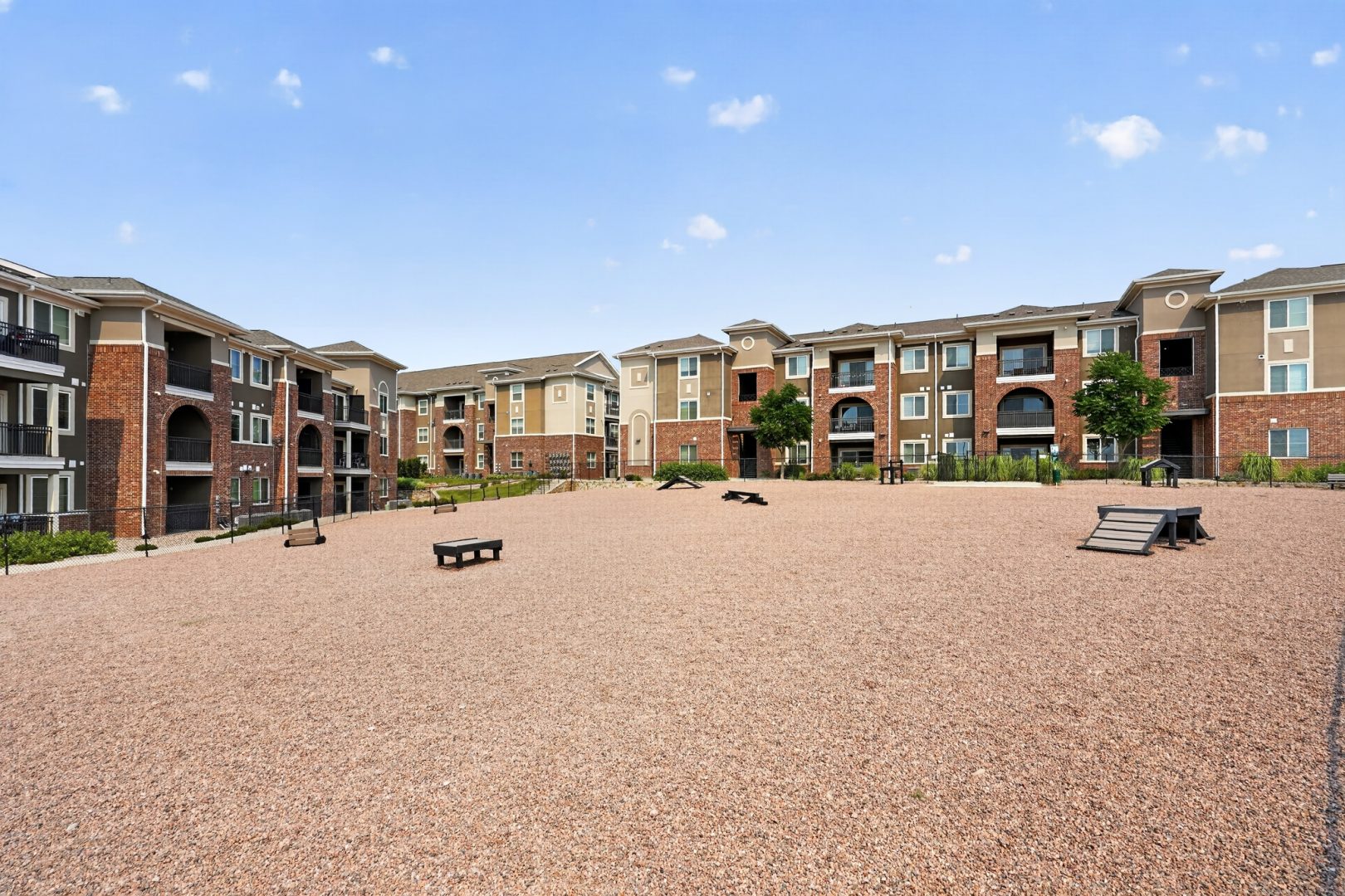 A gravel dog park with agility equipment, surrounded by multi-story apartment buildings featuring brick and balconies.
