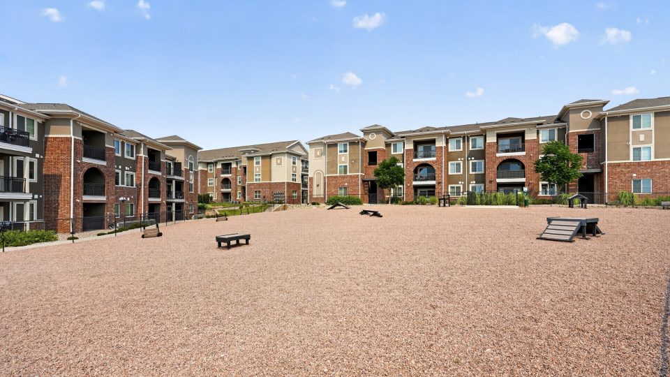 A gravel dog park with agility equipment, surrounded by multi-story apartment buildings featuring brick and balconies.