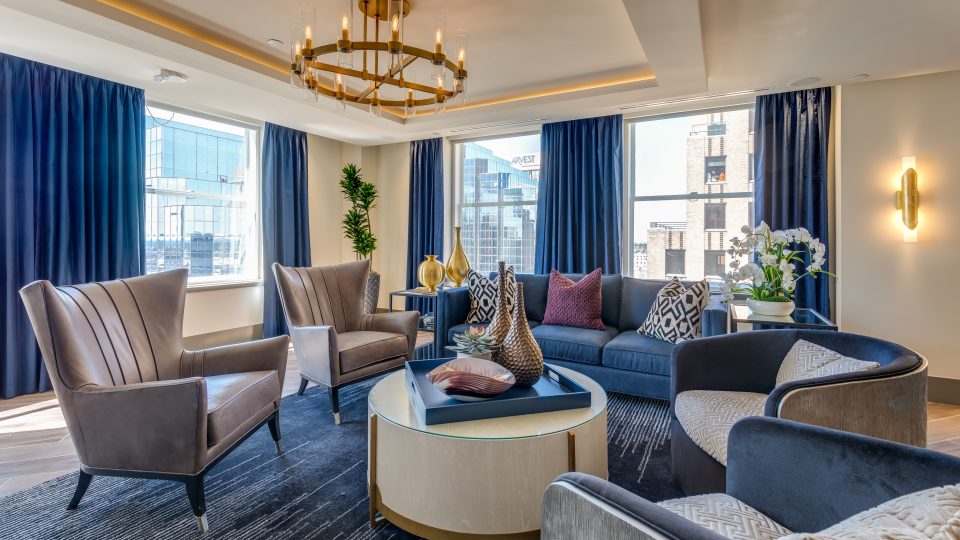 This InteriorPhoto shows a modern living room with a blue sofa, gray leather chairs, a round coffee table, and large windows with elegant navy curtains.