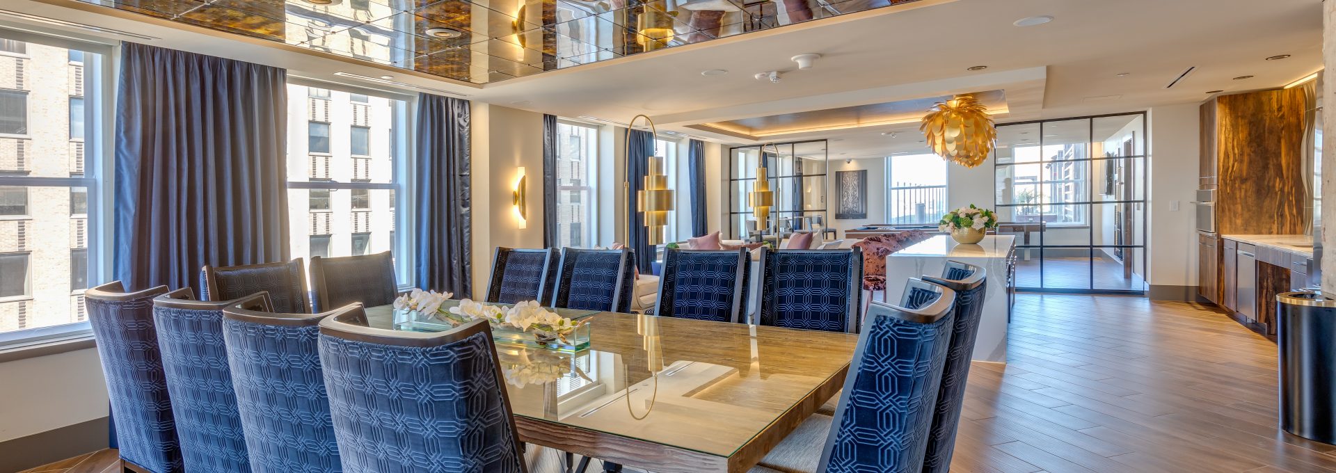 An interior photo of a dining area with a glass table and blue patterned chairs, a mirrored ceiling, and a modern open kitchen layout for a nice apartment.