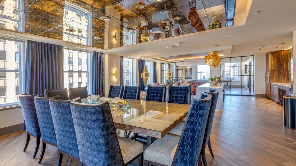 An interior photo of a dining area with a glass table and blue patterned chairs, a mirrored ceiling, and a modern open kitchen layout for a nice apartment.