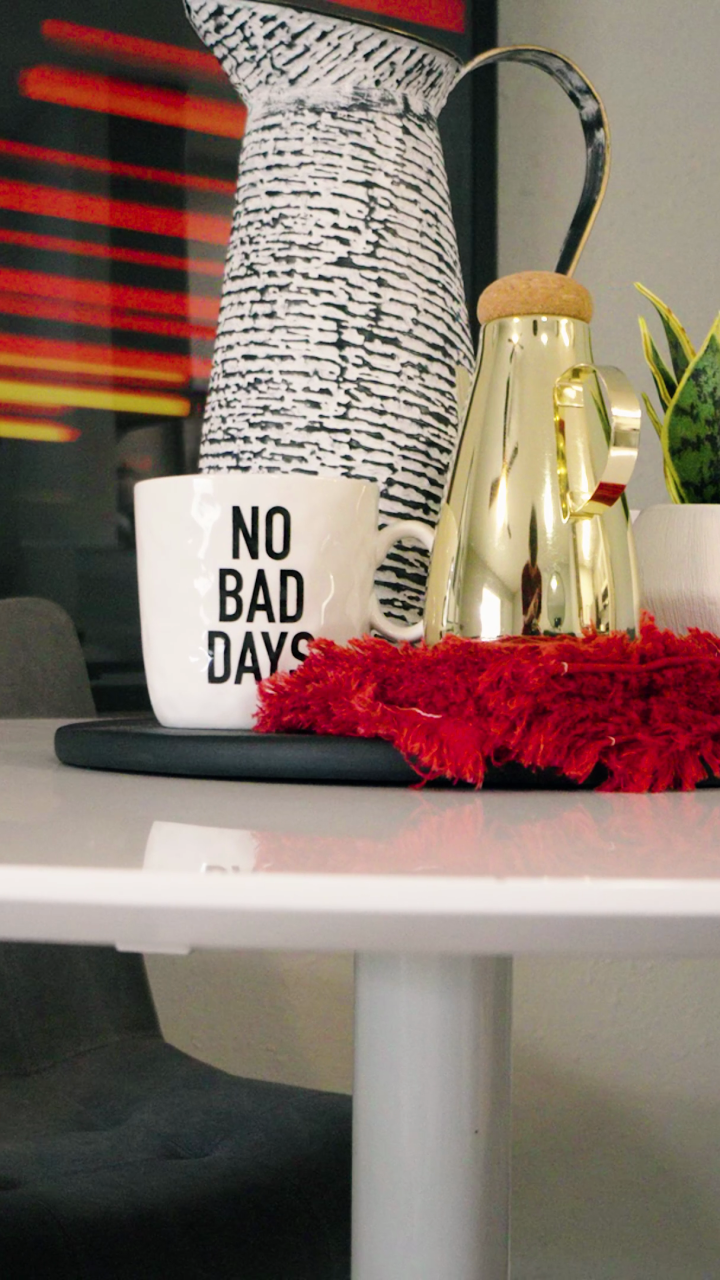 A cozy and stylish interior features a white table adorned with a gold vase, a small potted plant, and a mug that reads 'No Bad Days.' A red textured placemat adds a pop of color. The table is set against a vibrant, abstract painting, creating a lively and inviting atmosphere. The space is decorated with modern furniture, including a sleek, black chair, suggesting a comfortable and contemporary living area.