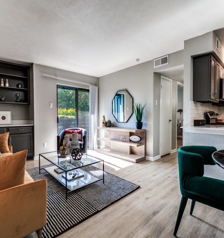 A cozy and modern living room features a comfortable orange sofa adorned with black and white checkered pillows. A glass coffee table sits atop a striped rug, adding a touch of elegance to the space. The room is illuminated by natural light streaming through a sliding glass door that opens to a balcony. A stylish dining area with green velvet chairs is adjacent to the kitchen, which boasts sleek black cabinets and stainless-steel appliances. The overall ambiance is inviting and perfect for both relaxation and socializing.