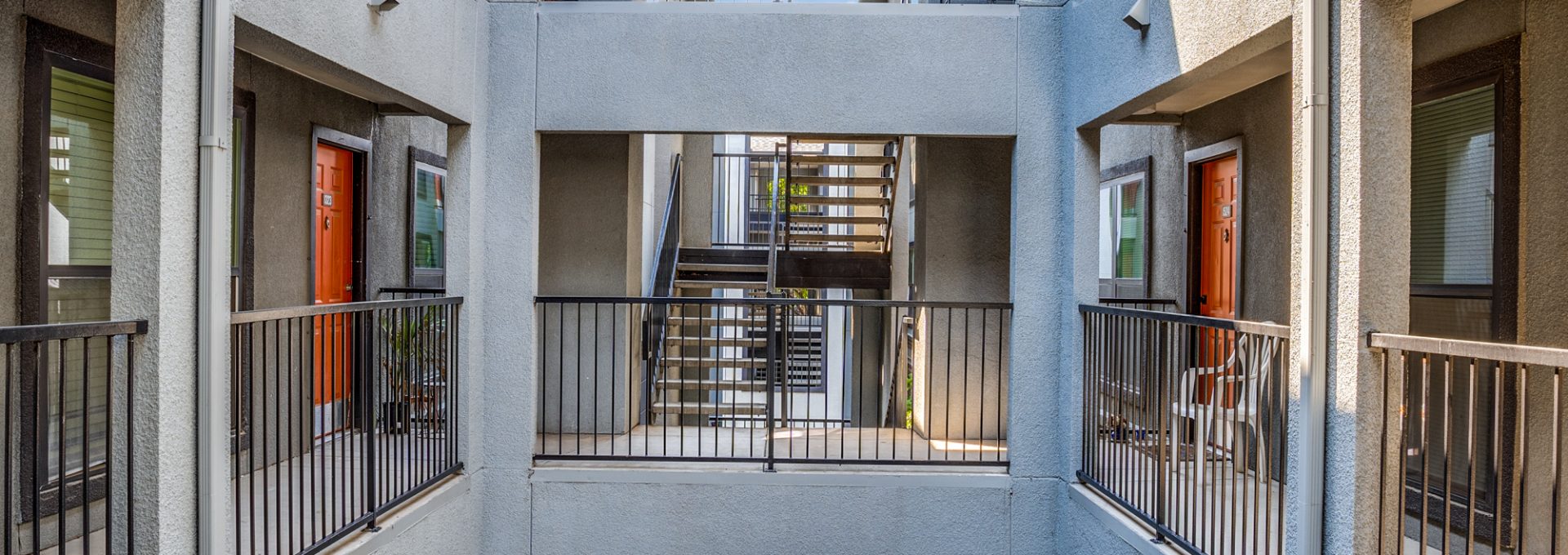 A quiet resident walkway passes through a bright, multi-level courtyard stairwell, using the open air above to move between floors. The primary activity appears to be navigating the building’s stair landing and corridor for everyday access. Sunlight streams through a skylight-like opening, highlighting sleek metal railings on both sides and textured stone walls. Concrete steps lead downward into a sheltered entry path, with a small outdoor mat at the lower level and a tidy gravel courtyard below that adds a calm, functional touch.