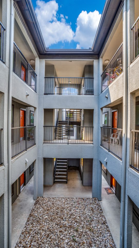A quiet resident walkway passes through a bright, multi-level courtyard stairwell, using the open air above to move between floors. The primary activity appears to be navigating the building’s stair landing and corridor for everyday access. Sunlight streams through a skylight-like opening, highlighting sleek metal railings on both sides and textured stone walls. Concrete steps lead downward into a sheltered entry path, with a small outdoor mat at the lower level and a tidy gravel courtyard below that adds a calm, functional touch.