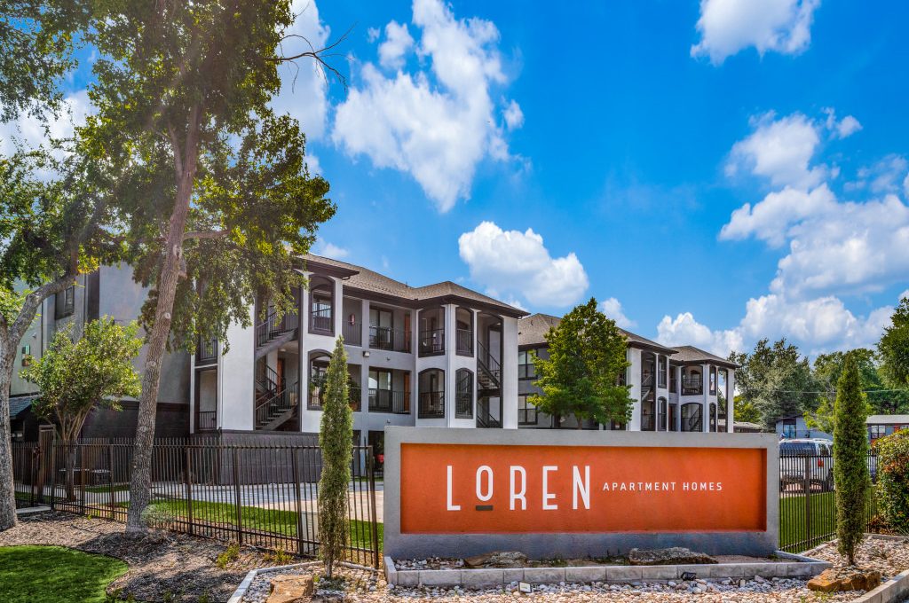 An outdoor apartment community courtyard features an inviting main entry sign reading “LQREN.” The primary activity is the welcoming arrival experience, guiding residents and visitors through a landscaped, resort-like space. Bright daylight illuminates modern two- to three-story building façades with arched windows and sleek balconies. A prominent orange sign panel sits atop a tidy stone base surrounded by gravel and river-rock landscaping. Neatly trimmed grass, decorative shrubs, and slender trees frame the walkway and fence line, creating a calm, well-maintained atmosphere for everyday living.