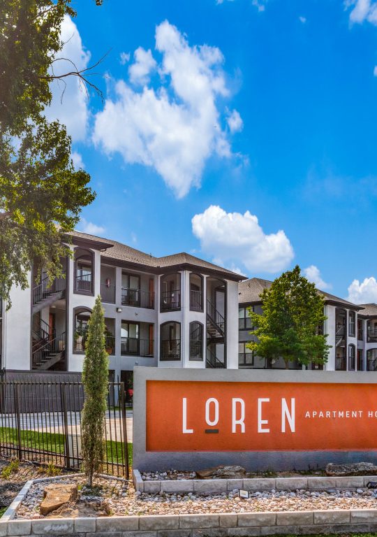 An outdoor apartment community courtyard features an inviting main entry sign reading “LQREN.” The primary activity is the welcoming arrival experience, guiding residents and visitors through a landscaped, resort-like space. Bright daylight illuminates modern two- to three-story building façades with arched windows and sleek balconies. A prominent orange sign panel sits atop a tidy stone base surrounded by gravel and river-rock landscaping. Neatly trimmed grass, decorative shrubs, and slender trees frame the walkway and fence line, creating a calm, well-maintained atmosphere for everyday living.