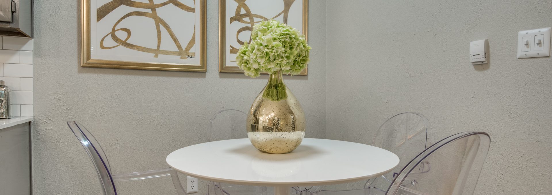 A stylish tabletop centerpiece takes center stage on a small round dining table, presenting a fresh bouquet in a shimmering vase. The cozy dining nook feels inviting and well-decorated, with clear acrylic chairs arranged neatly around the surface. Two framed abstract artworks and a pair of framed prints hang above, adding modern wall character. Soft neutral walls keep the space bright, while elegant glass and metal wall fixtures complement the clean, contemporary look. The arrangement suggests relaxed meals and everyday entertaining in a refined, minimalist setting.