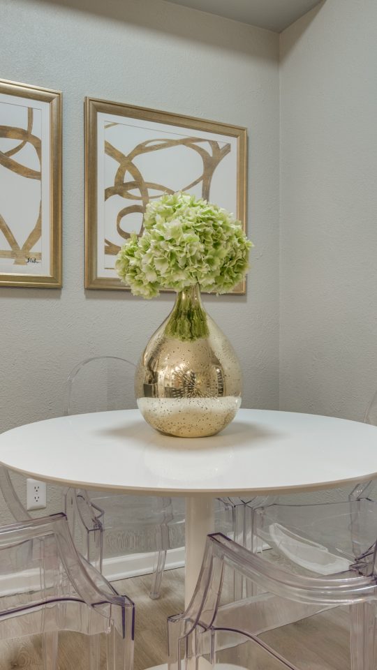 A stylish tabletop centerpiece takes center stage on a small round dining table, presenting a fresh bouquet in a shimmering vase. The cozy dining nook feels inviting and well-decorated, with clear acrylic chairs arranged neatly around the surface. Two framed abstract artworks and a pair of framed prints hang above, adding modern wall character. Soft neutral walls keep the space bright, while elegant glass and metal wall fixtures complement the clean, contemporary look. The arrangement suggests relaxed meals and everyday entertaining in a refined, minimalist setting.