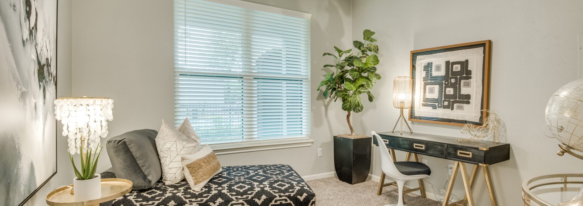 A person is not visible; the primary focus is a cozy reading and work nook where a comfortable sofa invites relaxing downtime. The space is styled with a patterned ottoman/bench, decorative pillows, and a bright ceiling fan overhead for comfortable everyday living. Large window blinds add soft, even light, while a small side table and a potted plant bring a fresh, welcoming touch. A sleek console with drawers holds a table lamp and framed wall art, and a white chair nearby creates a ready spot for writing or studying.