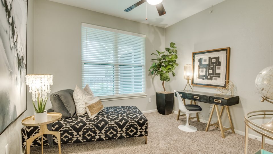 A person is not visible; the primary focus is a cozy reading and work nook where a comfortable sofa invites relaxing downtime. The space is styled with a patterned ottoman/bench, decorative pillows, and a bright ceiling fan overhead for comfortable everyday living. Large window blinds add soft, even light, while a small side table and a potted plant bring a fresh, welcoming touch. A sleek console with drawers holds a table lamp and framed wall art, and a white chair nearby creates a ready spot for writing or studying.