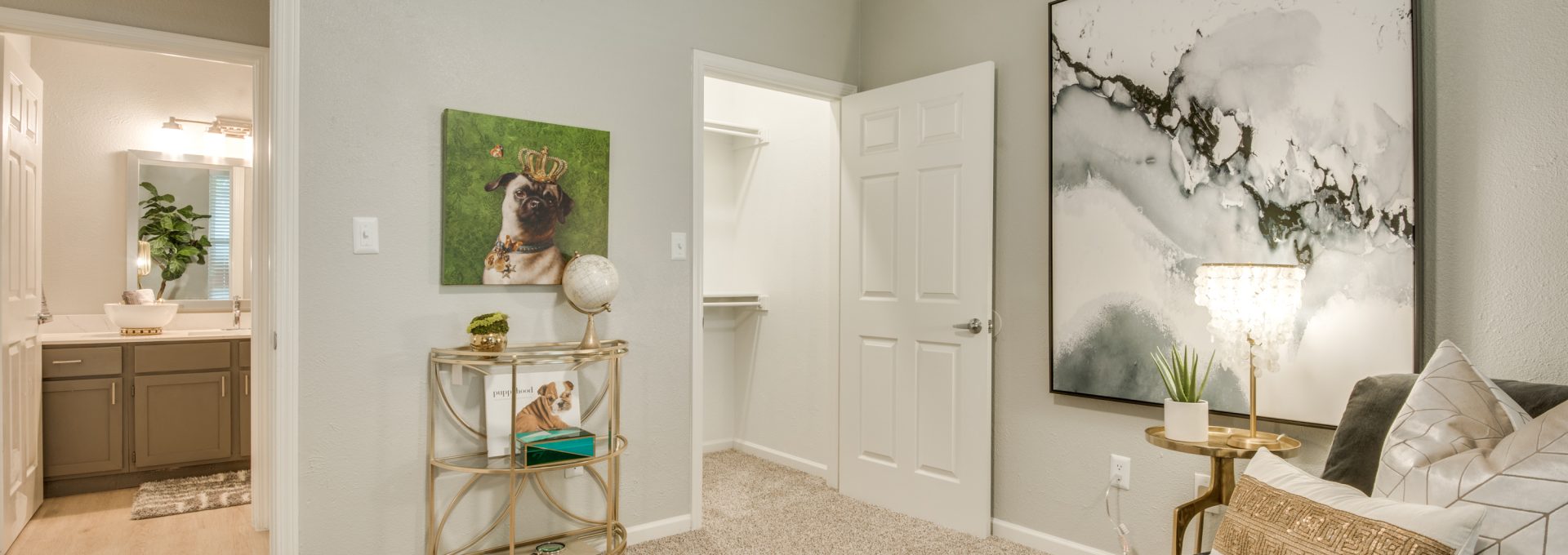A person relaxes on a sofa in a bright, stylish bedroom or lounge, enjoying a calm downtime moment. The space is arranged for comfort, featuring a neutral upholstered couch with patterned pillows and warm accent lighting from a modern ceiling fan. Gray carpet covers the floor, while framed wall art and a decorative table add a curated touch. Nearby, an open doorway reveals a bathroom vanity and plants, and a small round side table displays decor for an organized, homey feel.