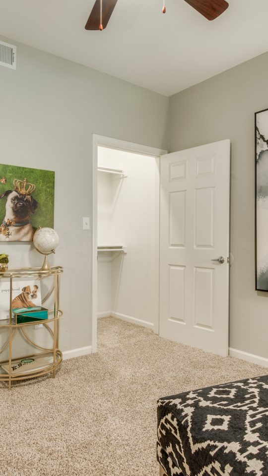 A person relaxes on a sofa in a bright, stylish bedroom or lounge, enjoying a calm downtime moment. The space is arranged for comfort, featuring a neutral upholstered couch with patterned pillows and warm accent lighting from a modern ceiling fan. Gray carpet covers the floor, while framed wall art and a decorative table add a curated touch. Nearby, an open doorway reveals a bathroom vanity and plants, and a small round side table displays decor for an organized, homey feel.