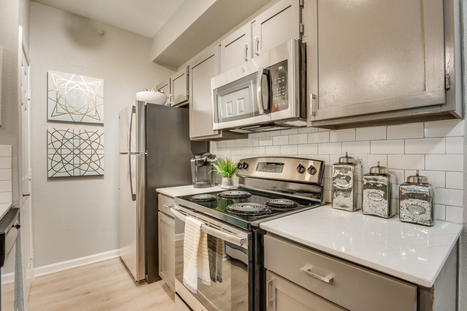 A staged kitchen features a stainless steel gas range as the focal point, with the stove top ready for cooking and meal prep. Bright white cabinets, a tiled backsplash, and sleek countertop surfaces create a clean, modern cooking space. The range is paired with matching stainless appliances, including a refrigerator and a built-in microwave above the cooktop. A bright under-cabinet hood and organized counter items such as a spice set and glass storage containers add a polished, lived-in feel while emphasizing everyday functionality and comfort.
