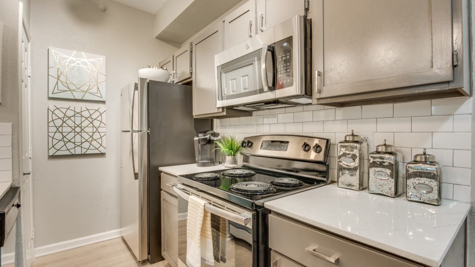 A staged kitchen features a stainless steel gas range as the focal point, with the stove top ready for cooking and meal prep. Bright white cabinets, a tiled backsplash, and sleek countertop surfaces create a clean, modern cooking space. The range is paired with matching stainless appliances, including a refrigerator and a built-in microwave above the cooktop. A bright under-cabinet hood and organized counter items such as a spice set and glass storage containers add a polished, lived-in feel while emphasizing everyday functionality and comfort.