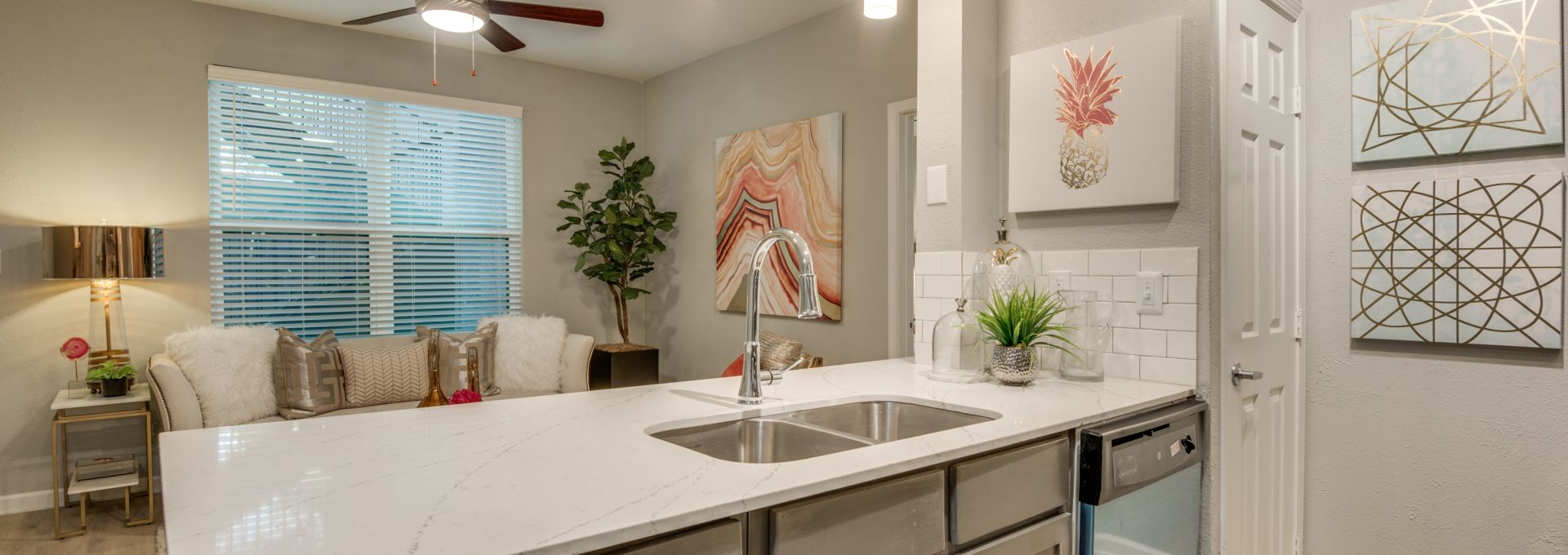 A bright, modern kitchen is shown from the countertop with the main activity centered on preparing and serving meals. The expansive white marble-look island features a built-in double sink and a sleek chrome faucet. Pendant lights with glowing shades hang overhead, complementing the clean, open layout. Under-cabinet storage and stainless dishwasher drawers flank the workspace, while a small countertop setup includes decor and a potted plant for an inviting touch. Stylish framed wall art and soft neutral finishes add warmth and a polished, comfortable feel.