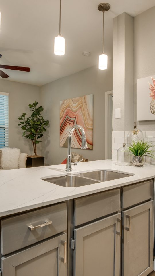 A bright, modern kitchen is shown from the countertop with the main activity centered on preparing and serving meals. The expansive white marble-look island features a built-in double sink and a sleek chrome faucet. Pendant lights with glowing shades hang overhead, complementing the clean, open layout. Under-cabinet storage and stainless dishwasher drawers flank the workspace, while a small countertop setup includes decor and a potted plant for an inviting touch. Stylish framed wall art and soft neutral finishes add warmth and a polished, comfortable feel.