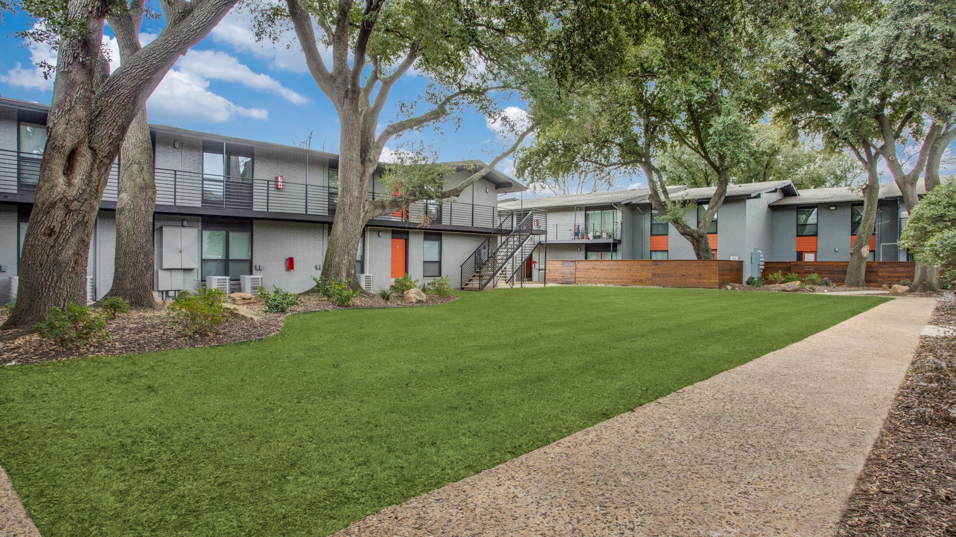 A sunny courtyard setting invites residents to enjoy outdoor living, with a well-kept green lawn in the foreground. The main activity is leisurely strolling along the walkway and relaxing in the landscaped common area. Mature trees cast welcoming shade over modern two-story apartments with sleek balconies and clean lines. A sturdy metal staircase connects the levels, while a low wood privacy screen and tidy garden beds frame the lawn, creating a calm, community feel.