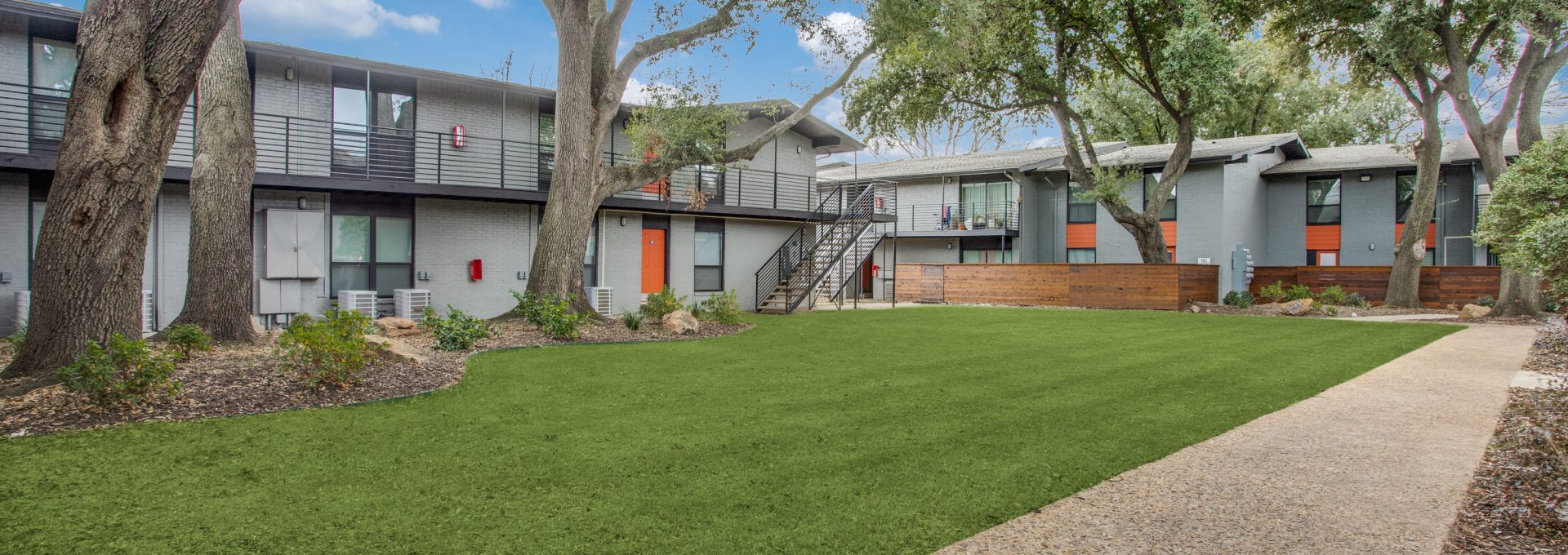 A sunny courtyard setting invites residents to enjoy outdoor living, with a well-kept green lawn in the foreground. The main activity is leisurely strolling along the walkway and relaxing in the landscaped common area. Mature trees cast welcoming shade over modern two-story apartments with sleek balconies and clean lines. A sturdy metal staircase connects the levels, while a low wood privacy screen and tidy garden beds frame the lawn, creating a calm, community feel.