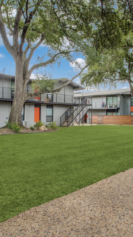 A sunny courtyard setting invites residents to enjoy outdoor living, with a well-kept green lawn in the foreground. The main activity is leisurely strolling along the walkway and relaxing in the landscaped common area. Mature trees cast welcoming shade over modern two-story apartments with sleek balconies and clean lines. A sturdy metal staircase connects the levels, while a low wood privacy screen and tidy garden beds frame the lawn, creating a calm, community feel.