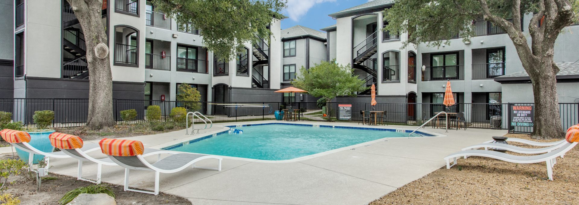 A bright outdoor swimming pool area invites residents to relax and soak up the sun. Lounge chairs with vibrant orange-and-white striped cushions sit beside the shimmering pool, while nearby patio tables offer a comfortable spot to unwind. The scene features a rectangular in-ground pool with a clean concrete deck, umbrella shade, and black metal safety fencing around the perimeter. Large mature trees provide natural canopy, enhancing the resort-like feel of the shared courtyard. 