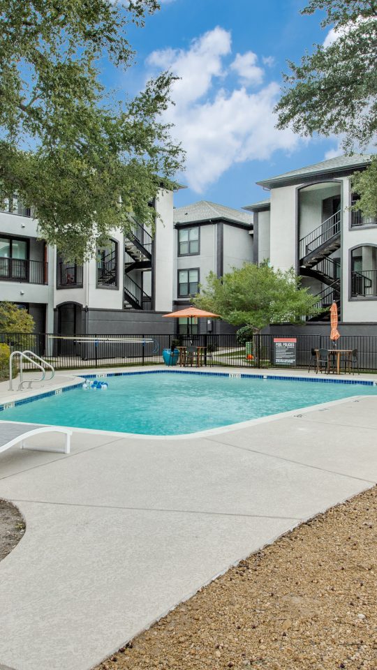 A bright outdoor swimming pool area invites residents to relax and soak up the sun. Lounge chairs with vibrant orange-and-white striped cushions sit beside the shimmering pool, while nearby patio tables offer a comfortable spot to unwind. The scene features a rectangular in-ground pool with a clean concrete deck, umbrella shade, and black metal safety fencing around the perimeter. Large mature trees provide natural canopy, enhancing the resort-like feel of the shared courtyard. 